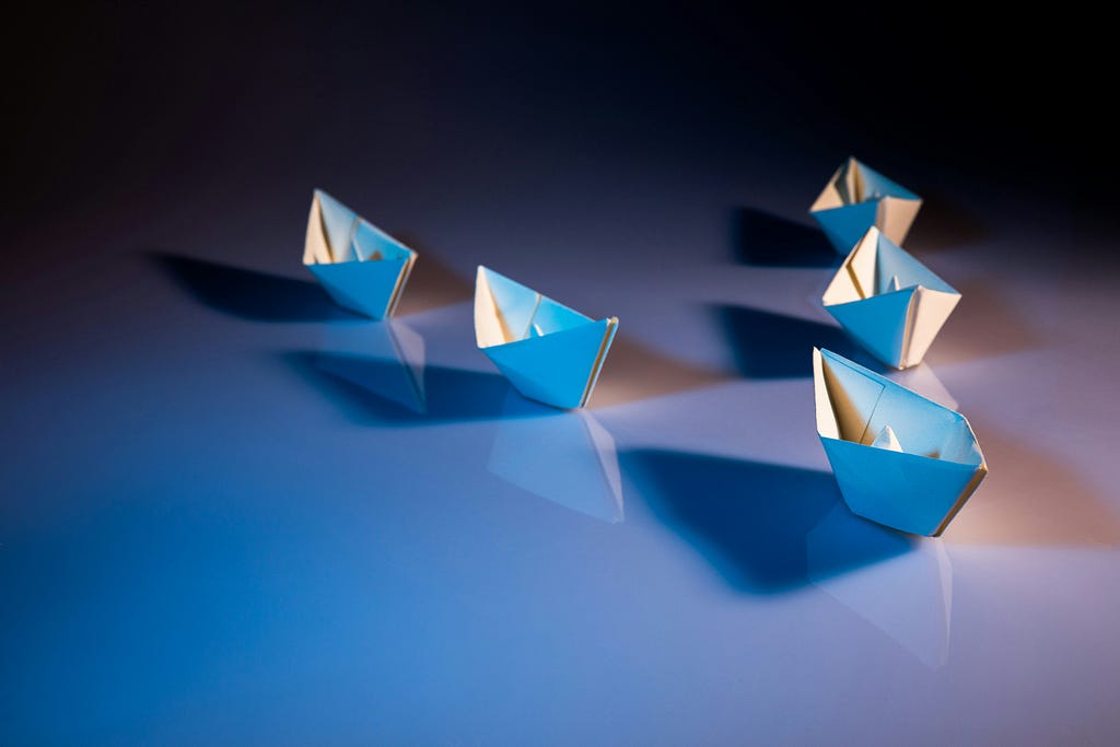 A group of paper boats arranged in a formation, with one boat leading the others. The boats are blue and white, and the scene is illuminated with soft lighting, creating shadows on a smooth surface.