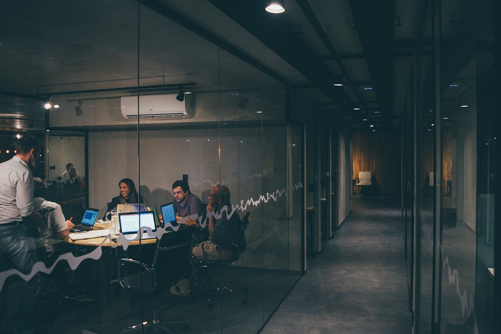 A group of people working late in a modern office meeting room, with glass walls and dim lighting. The team is engaged in discussion and using laptops, creating a focused and collaborative atmosphere.