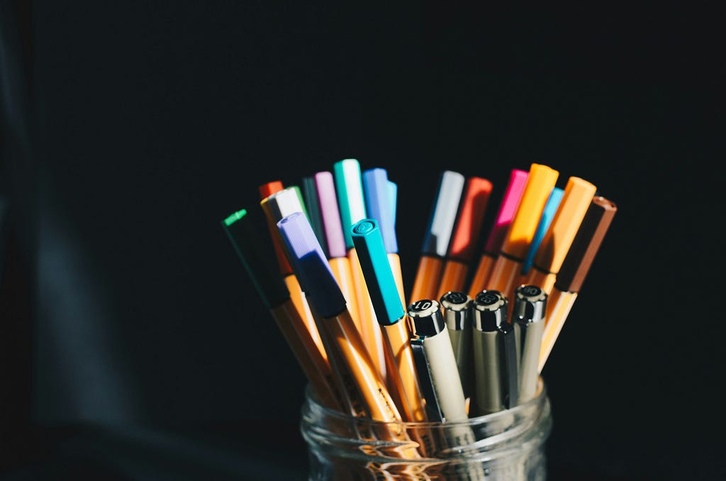 A glass jar filled with colorful markers and pens, illuminated by light against a dark background.