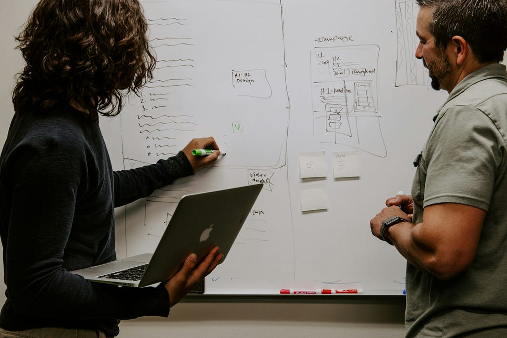 Two people collaborating at a whiteboard, one holding a laptop and writing with a marker, while the other observes and discusses the sketches and notes on the board.