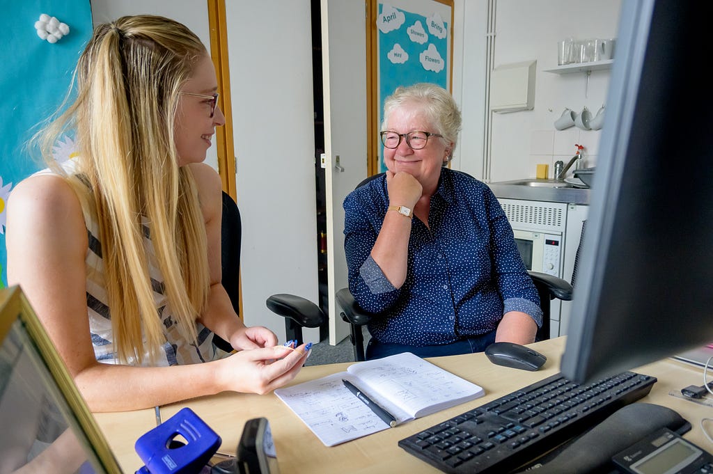 Young woman talking to an older woman sat at a desktop computer in an office/study room at home.